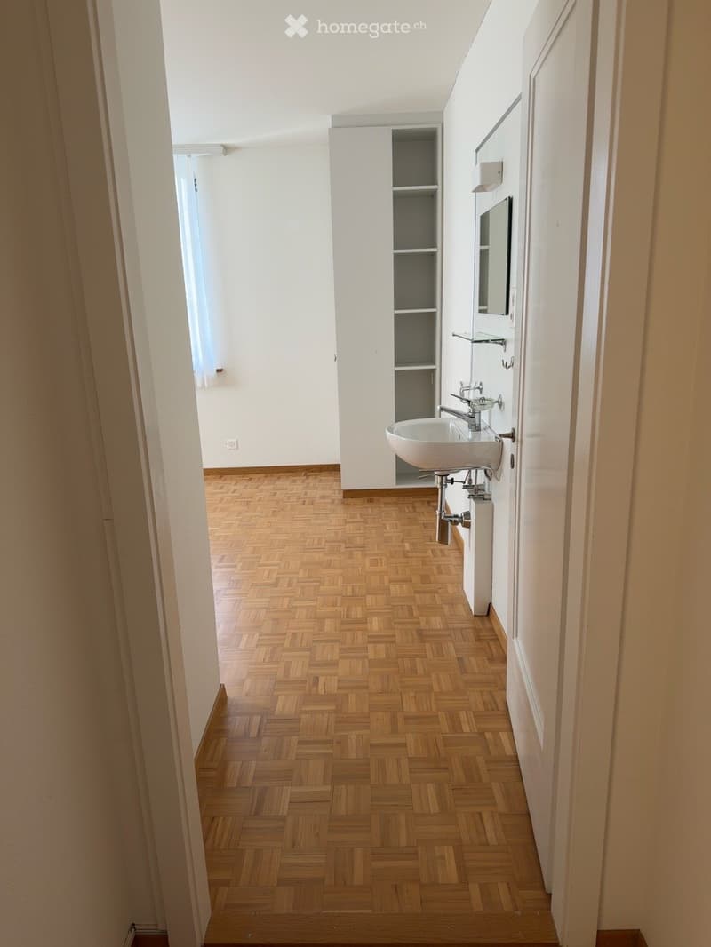 A narrow hallway with wooden herringbone flooring, white walls, and a small sink area visible at the end.