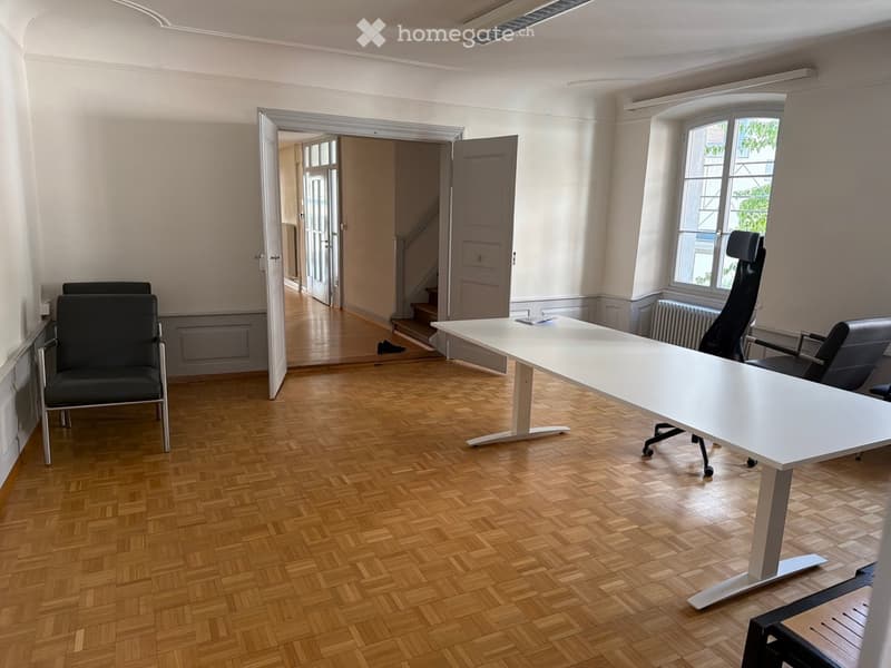 Office space with wooden herringbone flooring, white table, black office chairs, large window with view, open doorway leading to hallway.