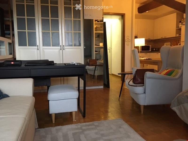 Bright, modern living space with hardwood flooring, a grey sofa, a small black desk with a laptop, a white armchair, a small round table, and a kitchen area with cabinets and a microwave.