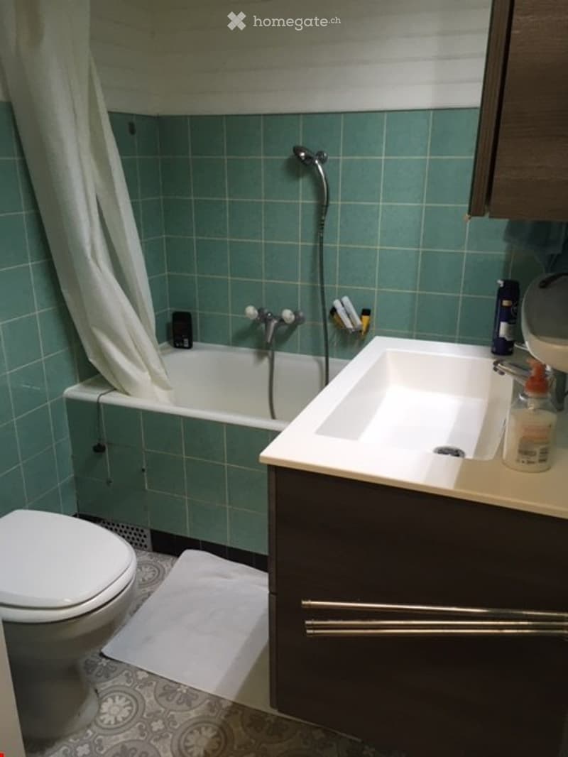 Bathroom with green tiled walls, a white bathtub, a toilet, a sink with a faucet, a showerhead, a shower curtain, and a cabinet.