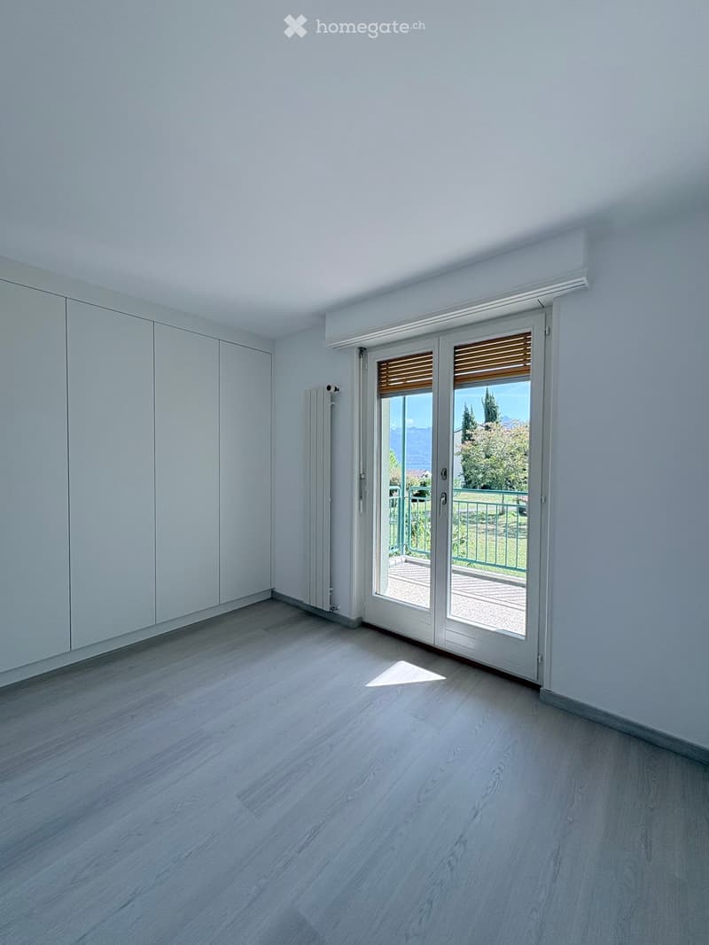 Empty room with light wooden flooring, white walls, large sliding glass doors leading to a balcony, and a radiator on one wall.