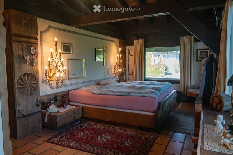 Cozy bedroom with wooden beams, a large bed with pink bedding, nightstand with a telephone, wall-mounted chandelier, framed artwork, and a large window with curtains.