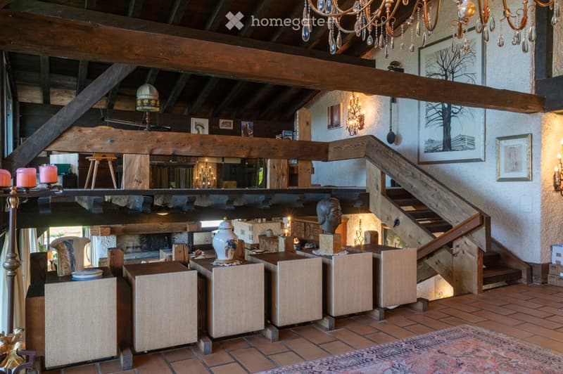 A spacious, well-lit living area with exposed wooden beams, a large staircase, and a cozy seating arrangement. The room features a mix of modern and rustic decor, including a large chandelier, wall-mounted lamps, and various decorative items on tables and