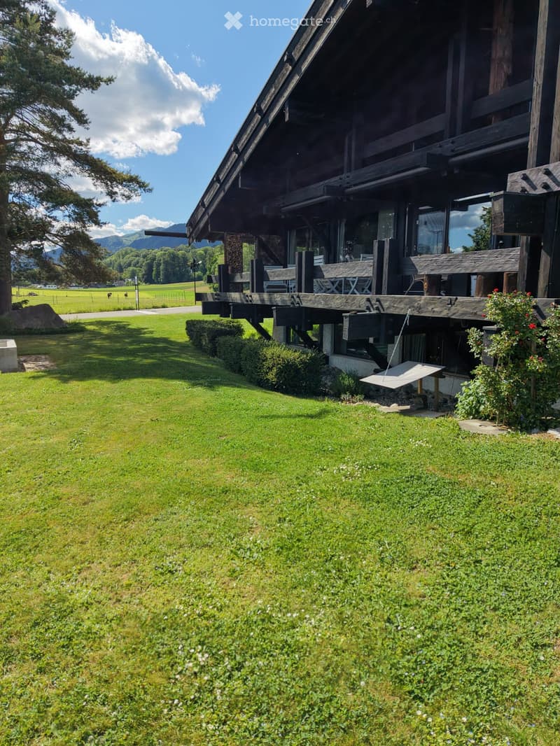 A large wooden house with a spacious wooden deck, surrounded by a well-maintained lawn and garden, with a scenic mountain view in the background.