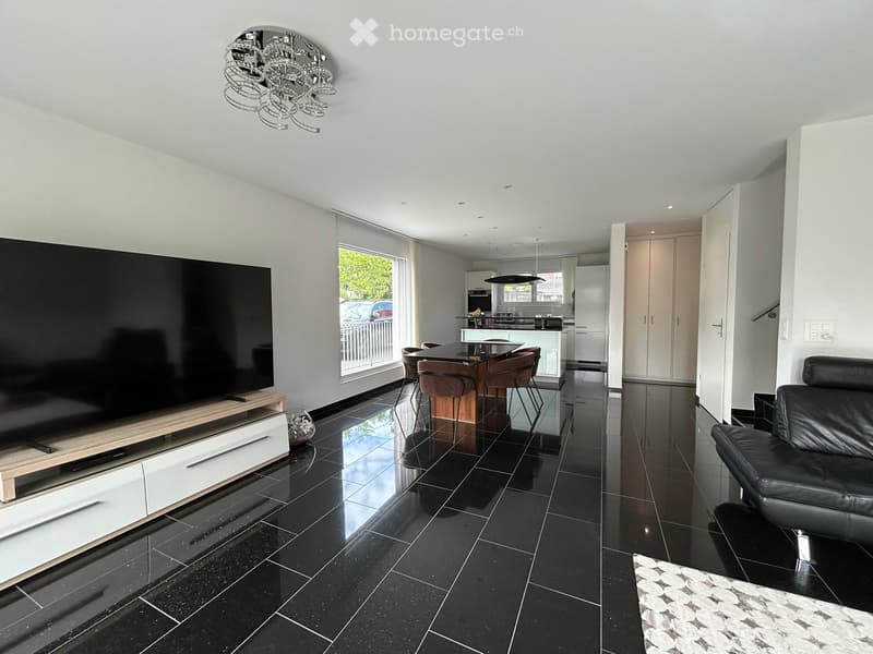 Modern living room with black tiled flooring, large flat-screen TV on a wooden stand, leather sofa, coffee table, dining area with a table and chairs, and an open kitchen area with cabinets and a kitchen island.