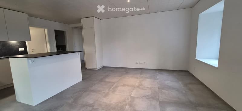 Empty kitchen with white cabinets, a black countertop, and a large window letting in natural light. The floor is tiled, and the space is clean and uncluttered.