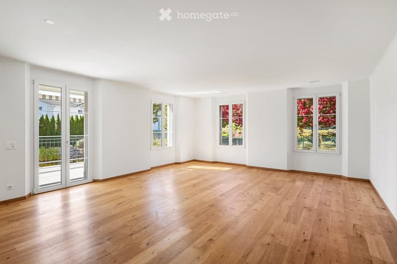 Empty room with light wooden flooring, white walls, and large windows with views of greenery outside. The space is unfurnished and well-lit.
