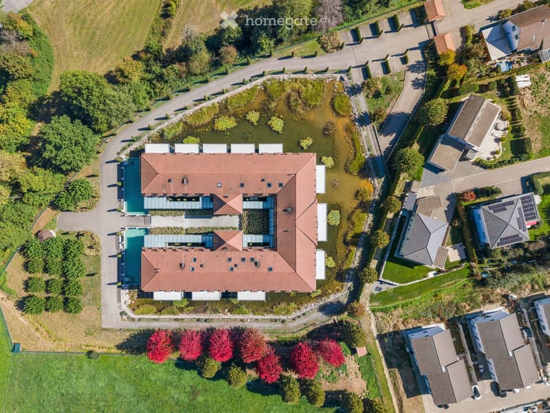 Aerial view of a large, modern house with a red-tiled roof, surrounded by a landscaped garden with a pond, pathways, and neighboring houses.