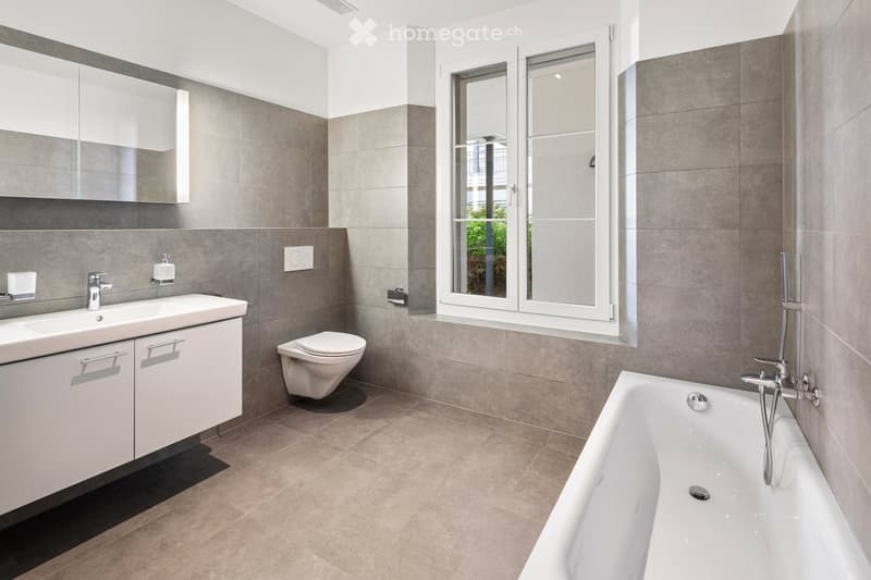 Modern bathroom with a bathtub, toilet, sink, and mirrored cabinet. Features grey tiles, a large window, and sleek fixtures.