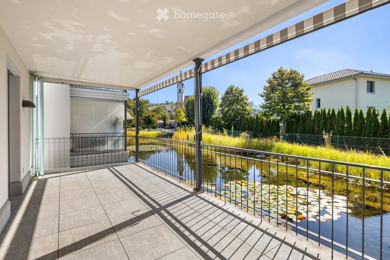 Balcony with a modern design, featuring a tiled floor, metal railing, and a view of a pond with lily pads. The balcony is covered by a retractable awning and overlooks a landscaped area with trees and a distant church tower.