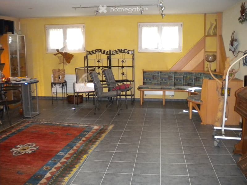 Brightly colored living room with yellow walls, tiled floor, and large windows. Features a sofa, chairs, a bench, a table, and decorative shelves.