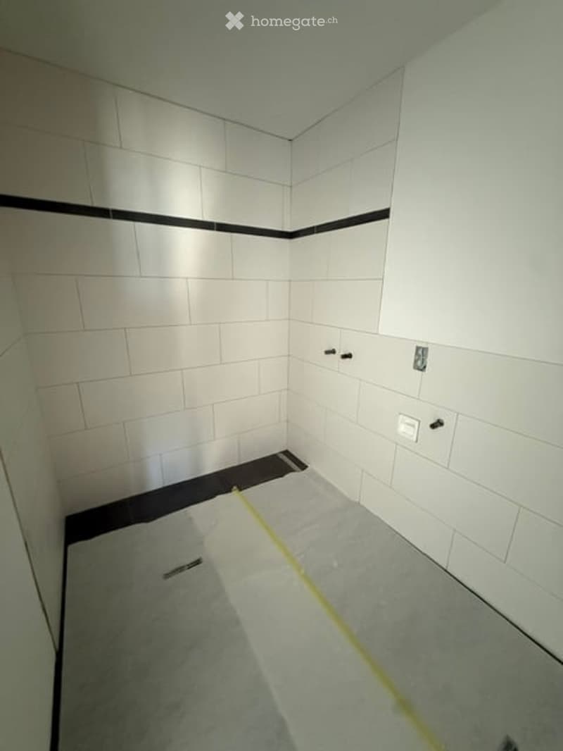 Empty bathroom with white tiled walls and floor, featuring a black border along the upper wall and a gray floor. The space appears to be under renovation or construction, with no fixtures or furniture present.