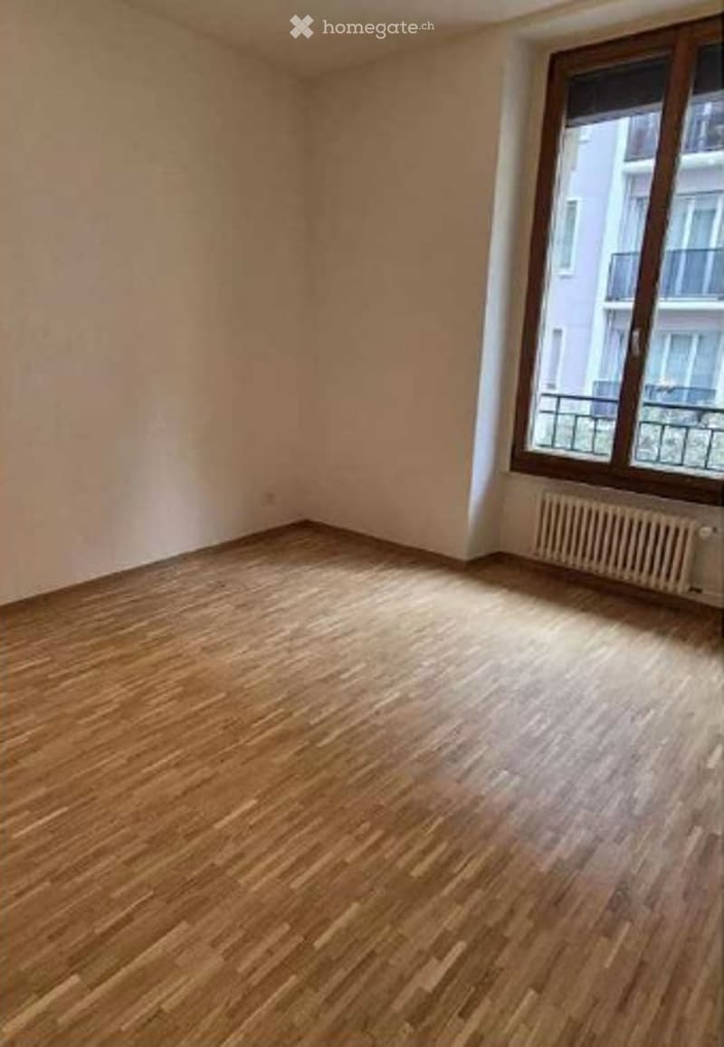 Empty room with wooden flooring, white walls, and a large window with a radiator below it.