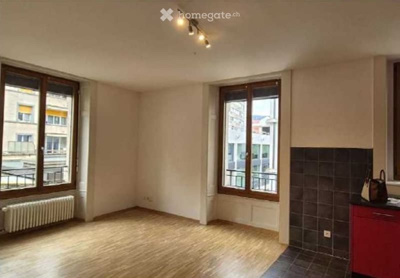 Empty room with wooden flooring, large windows, radiator, and a small red cabinet in the corner.