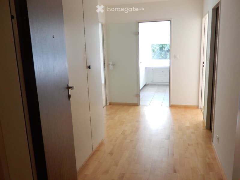 Empty hallway with light wooden flooring, white walls, and a glass door leading to a balcony area.
