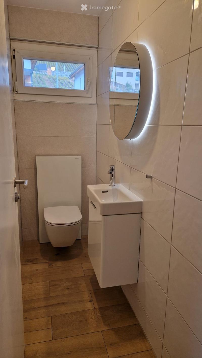 Modern bathroom with a toilet, sink, and mirror. The sink has a sleek, contemporary design with a chrome faucet, and the mirror is illuminated with soft lighting. The floor is made of light wooden planks, and the walls are tiled in a neutral tone.