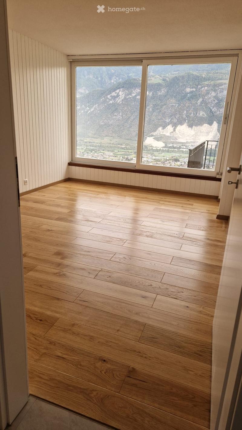 Empty room with wooden flooring, large windows offering a scenic mountain view, and white paneled walls.