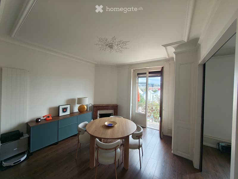 Bright, airy room with wooden flooring, a round wooden dining table with four chairs, a blue cabinet with a lamp, a framed picture, and a fireplace. A large window with a view opens to a balcony.