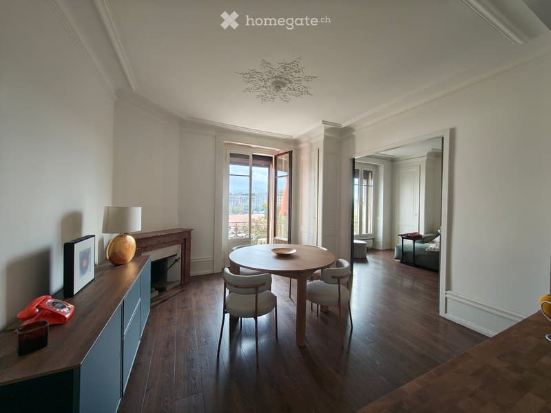 Elegant living and dining area with wooden flooring, a round dining table with chairs, a sideboard with a lamp and picture frame, and a view through large windows.