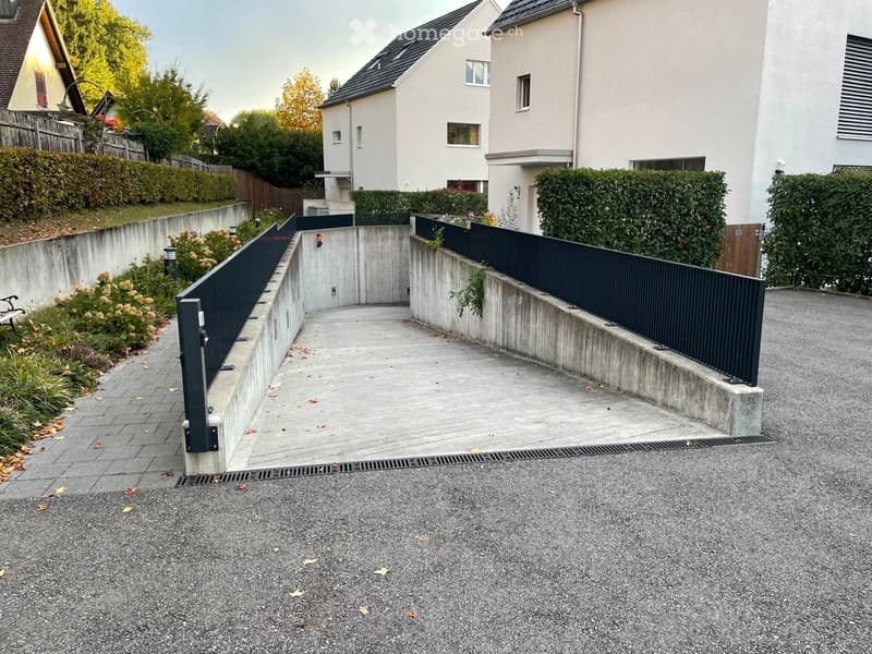 Driveway with concrete barriers, black metal fence, and surrounding greenery.