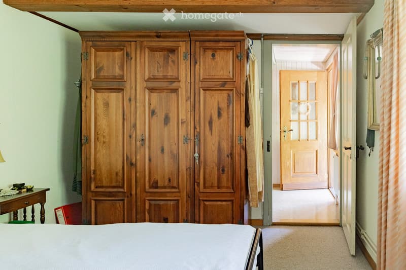 Cozy bedroom with a large wooden wardrobe, a neatly made bed, a small wooden table with a lamp, and a mirror on the wall. The room features light-colored walls and carpeted flooring, with a doorway leading to another room.