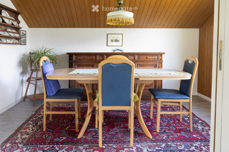 Dining room with a wooden table, four chairs with blue cushions, a wooden cabinet, a potted plant, and a chandelier.
