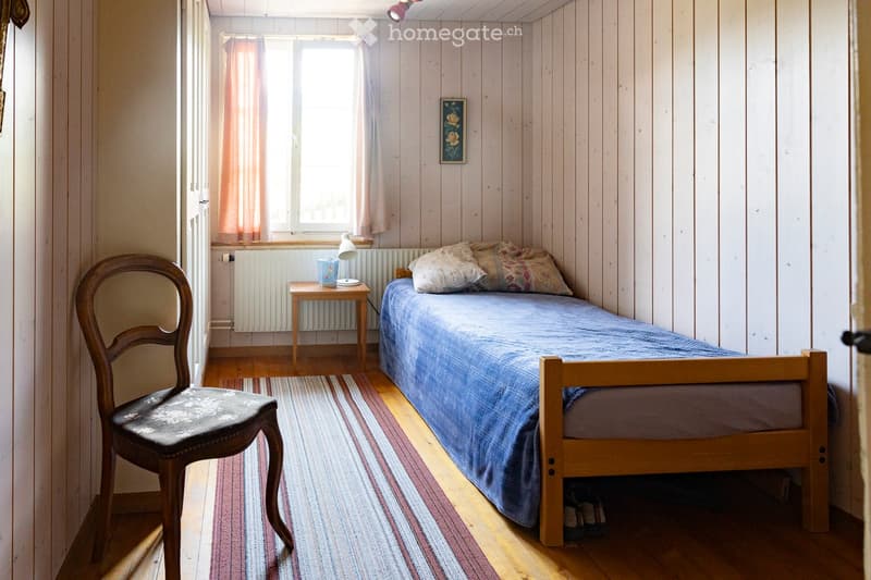 Cozy bedroom with wooden walls, a single bed with blue bedding, a wooden chair, a small table with a lamp, and a radiator under the window.