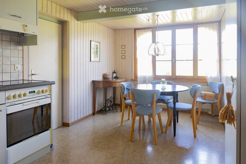 Bright kitchen with wooden panel walls, a dining area with a round table and six chairs, a stove, and a chandelier hanging from the ceiling.