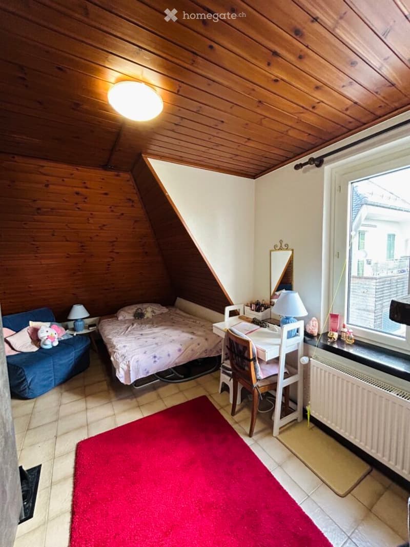 Cozy attic bedroom with wooden slanted ceilings, a bed with pink bedding, a small sofa, a desk with a chair, a mirror, and a red rug on the floor.