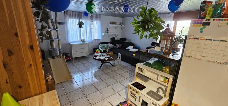 Living room with a black sofa, small round table, white cabinet, cat tree, and a toy kitchen set. There are balloons, plants, and a calendar on the refrigerator.