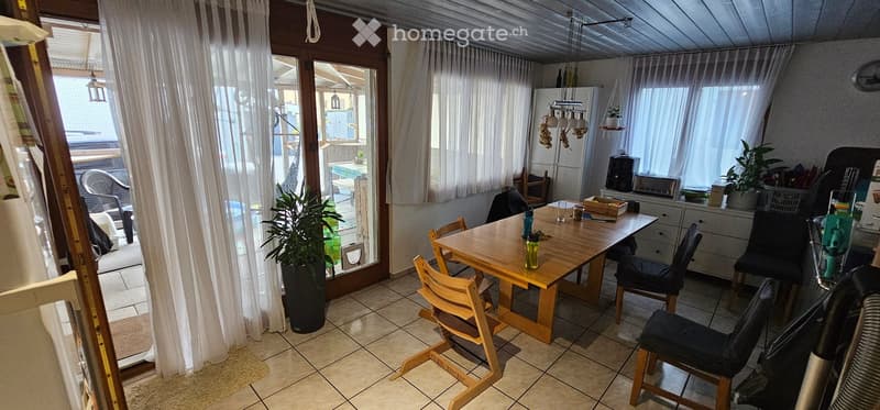 A well-lit dining area with a wooden table and chairs, a refrigerator, and a curtained window leading to a patio. There are plants and a small cabinet visible.
