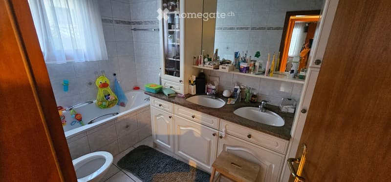 Bathroom with a bathtub, double sink, mirror, and various bathroom accessories and products. Includes a small stool and a toilet.