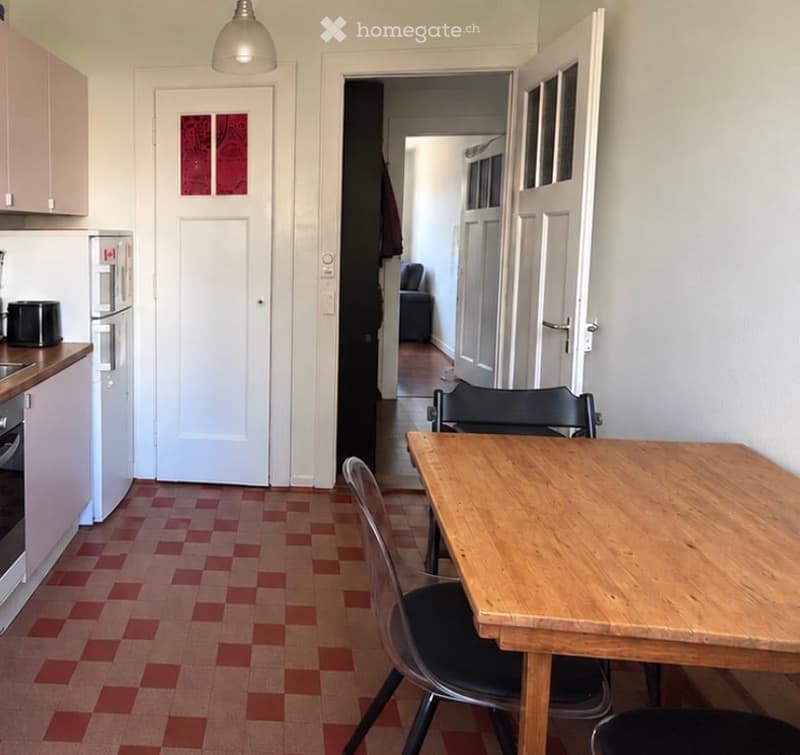 A small, well-lit kitchen and dining area with a wooden dining table, chairs, and modern appliances including a refrigerator and oven. The floor has a red and beige checkered pattern.