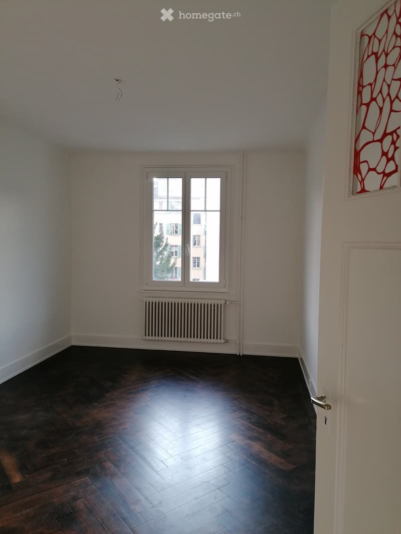 Empty room with wooden flooring, white walls, a window with white frame, and a radiator below the window.