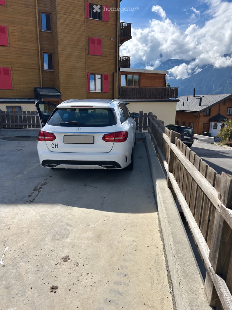 A parking area with a white Mercedes-Benz car, wooden fence, and a multi-story wooden building with red shutters and balconies in the background.