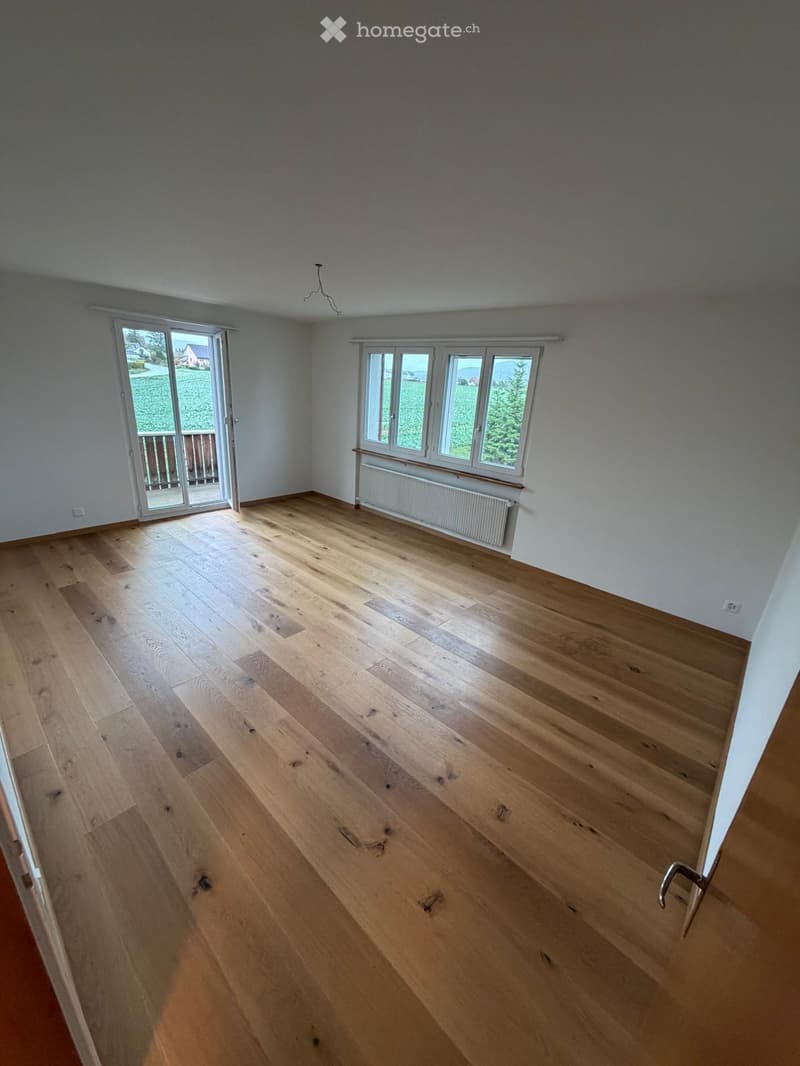Empty room with wooden flooring, large windows, and a sliding glass door leading to a balcony.