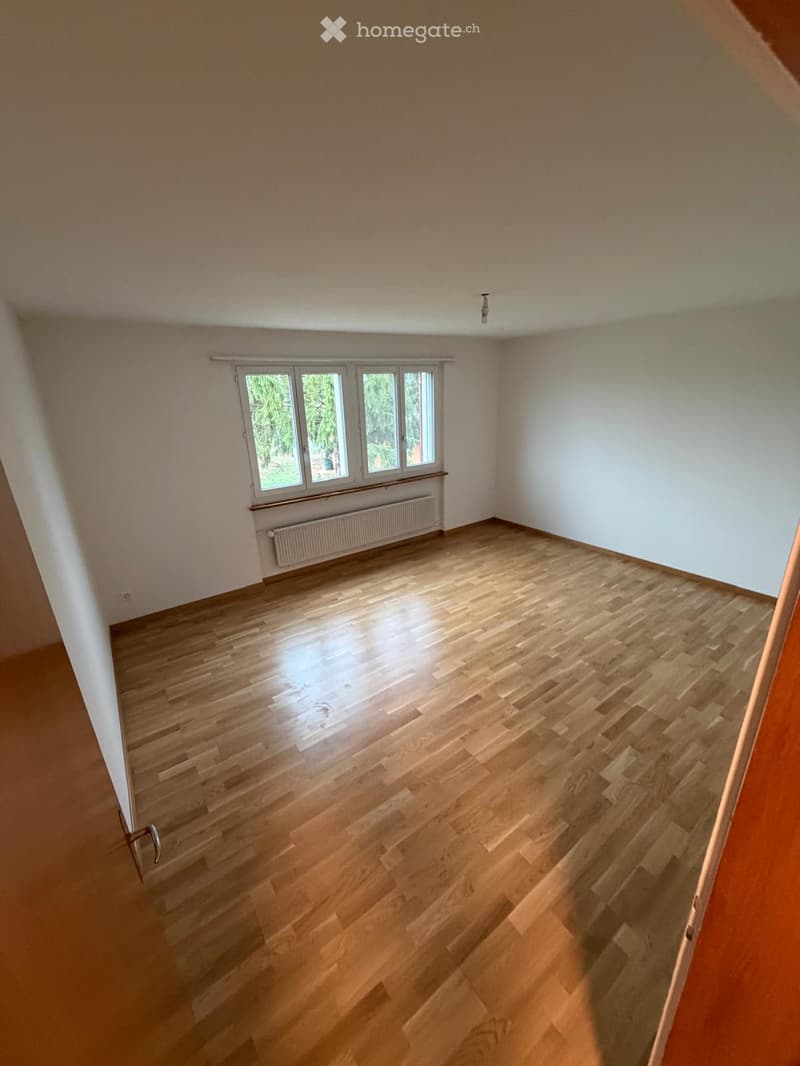 Empty room with wooden flooring, white walls, and large windows with a radiator below them.