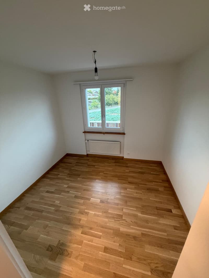 Empty room with wooden flooring, white walls, a window with a view of greenery, a radiator, and a ceiling light fixture.