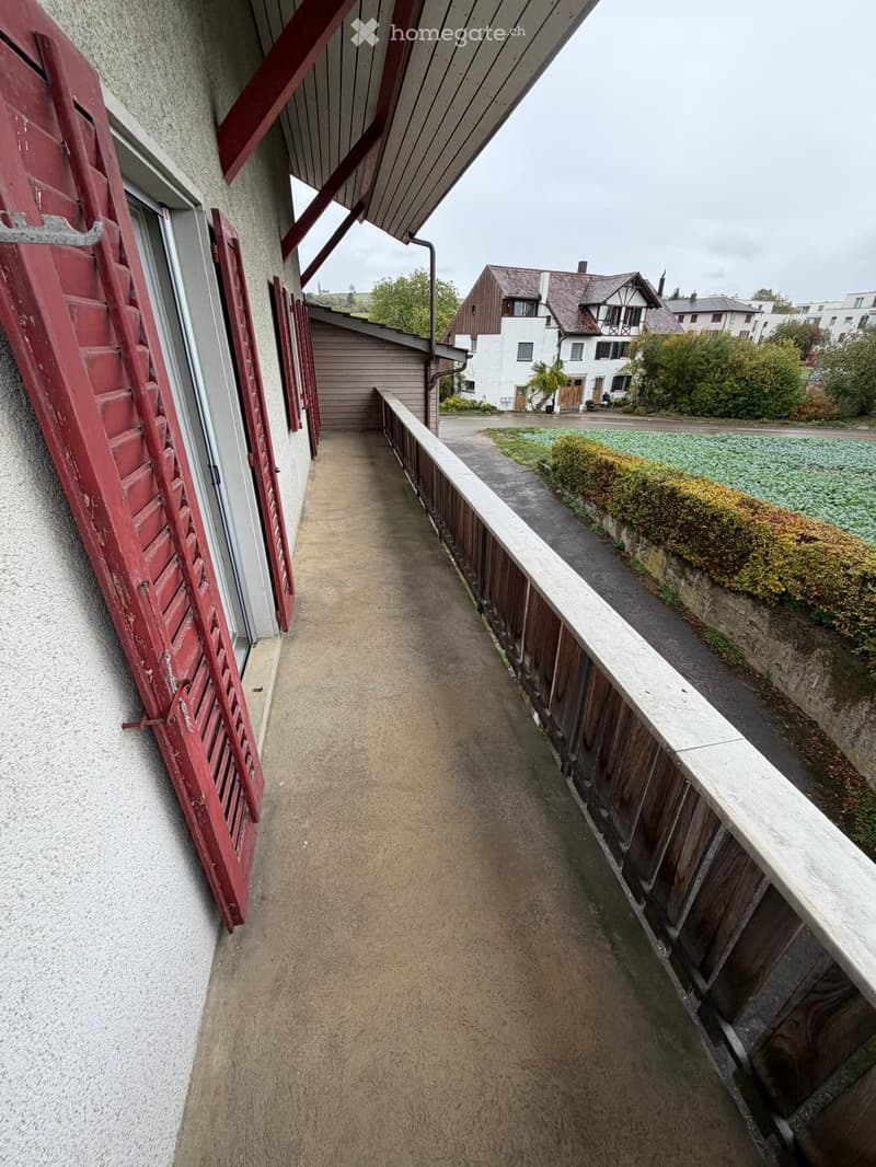 A narrow concrete pathway bordered by a wooden railing on one side and a building wall with red shutters on the other, leading to a residential area with houses and greenery.