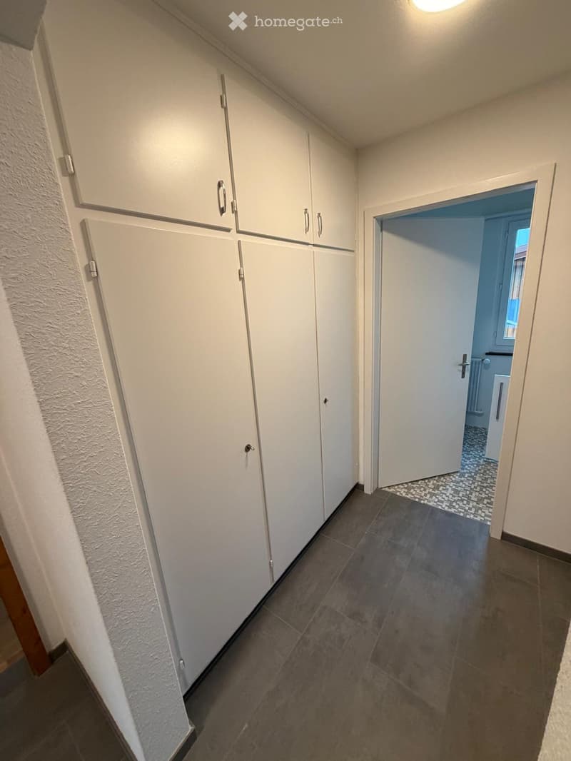 A narrow hallway with white built-in cabinets on one side and a tiled floor. An open doorway leads to a blue-tiled bathroom area visible in the background.