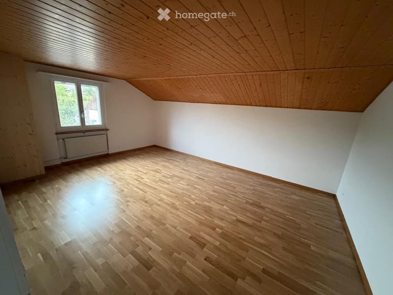 Empty attic room with wooden floor, white walls, and a slanted wooden ceiling. Features a single window and a radiator under the window.