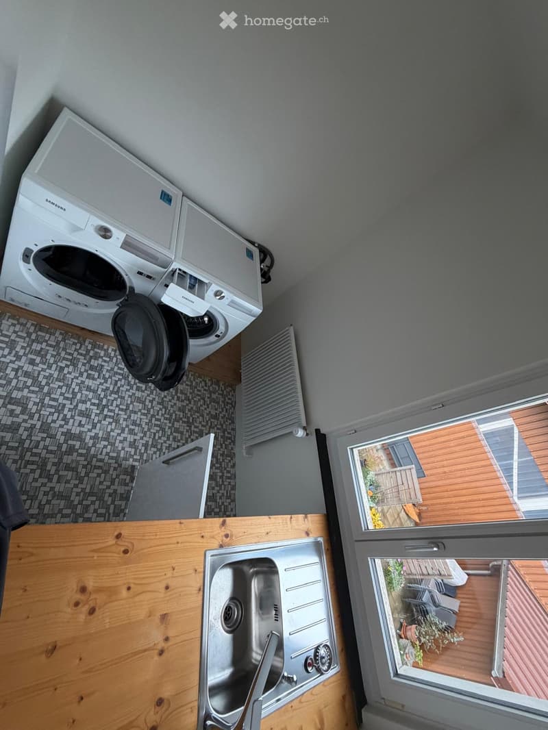 Bathroom with a stainless steel sink, wooden countertop, washing machine, radiator, and a window overlooking a wooden deck with outdoor furniture.