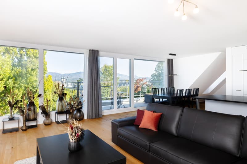 Modern living room with large windows offering a scenic view, featuring a black leather sofa, coffee table, and decorative vases.