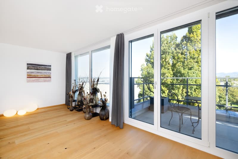 Bright living room with wooden flooring, large windows, and a balcony overlooking greenery. Decor includes vases, a chair, and a small table on the balcony.