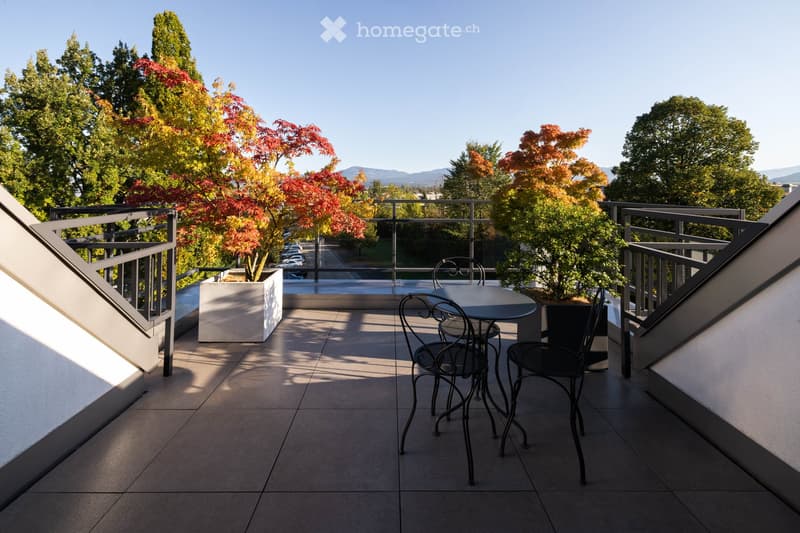 Balcony with a round table and four black metal chairs, surrounded by trees and a clear view of the street.