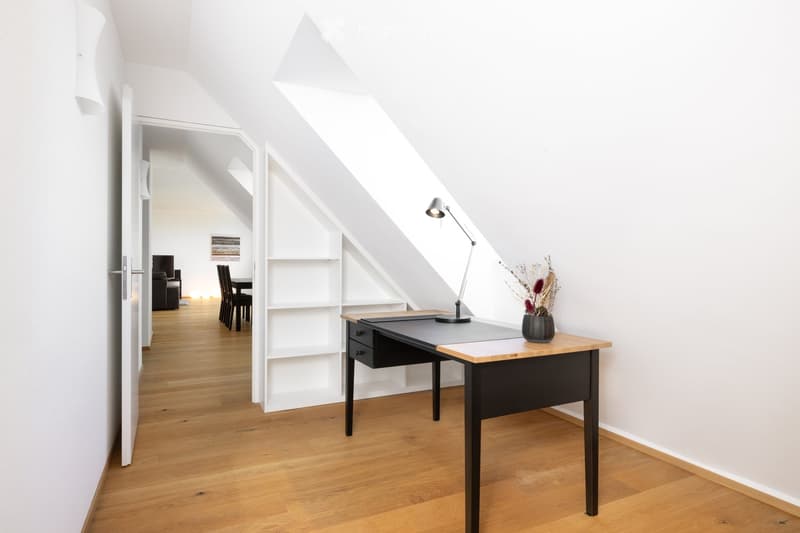Bright, modern attic room with wooden flooring, slanted ceilings, and built-in white shelving. Features a black desk with a lamp, a small table with a vase of dried flowers, and a dining area visible through an open doorway.