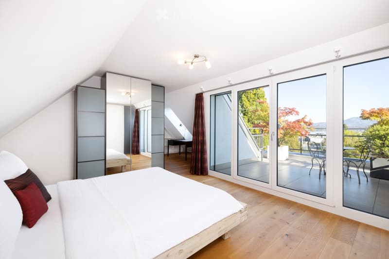 Bright bedroom with wooden flooring, large sliding glass doors leading to a balcony, a bed with white linens and red pillows, a mirrored wardrobe, a small table and chair, and a view of greenery and mountains.