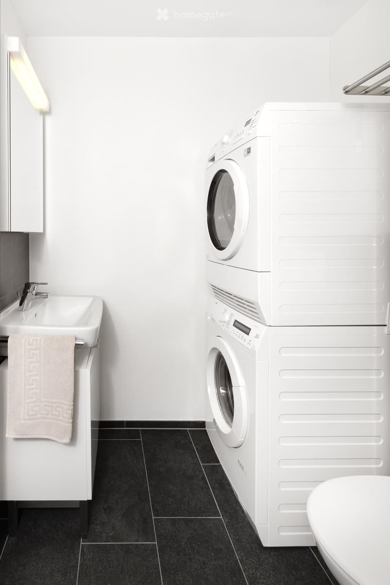 Modern bathroom with white walls, black tiled floor, white sink with faucet, towel, washing machine, and toilet.