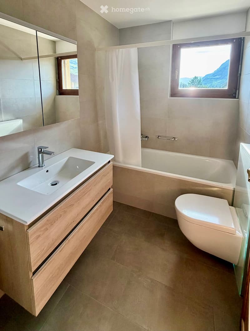 Modern bathroom with a white sink, wooden cabinet, bathtub, toilet, and large windows offering a scenic mountain view.