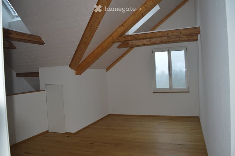 Empty room with wooden floor, white walls, exposed wooden beams, and a window with double glass panels.